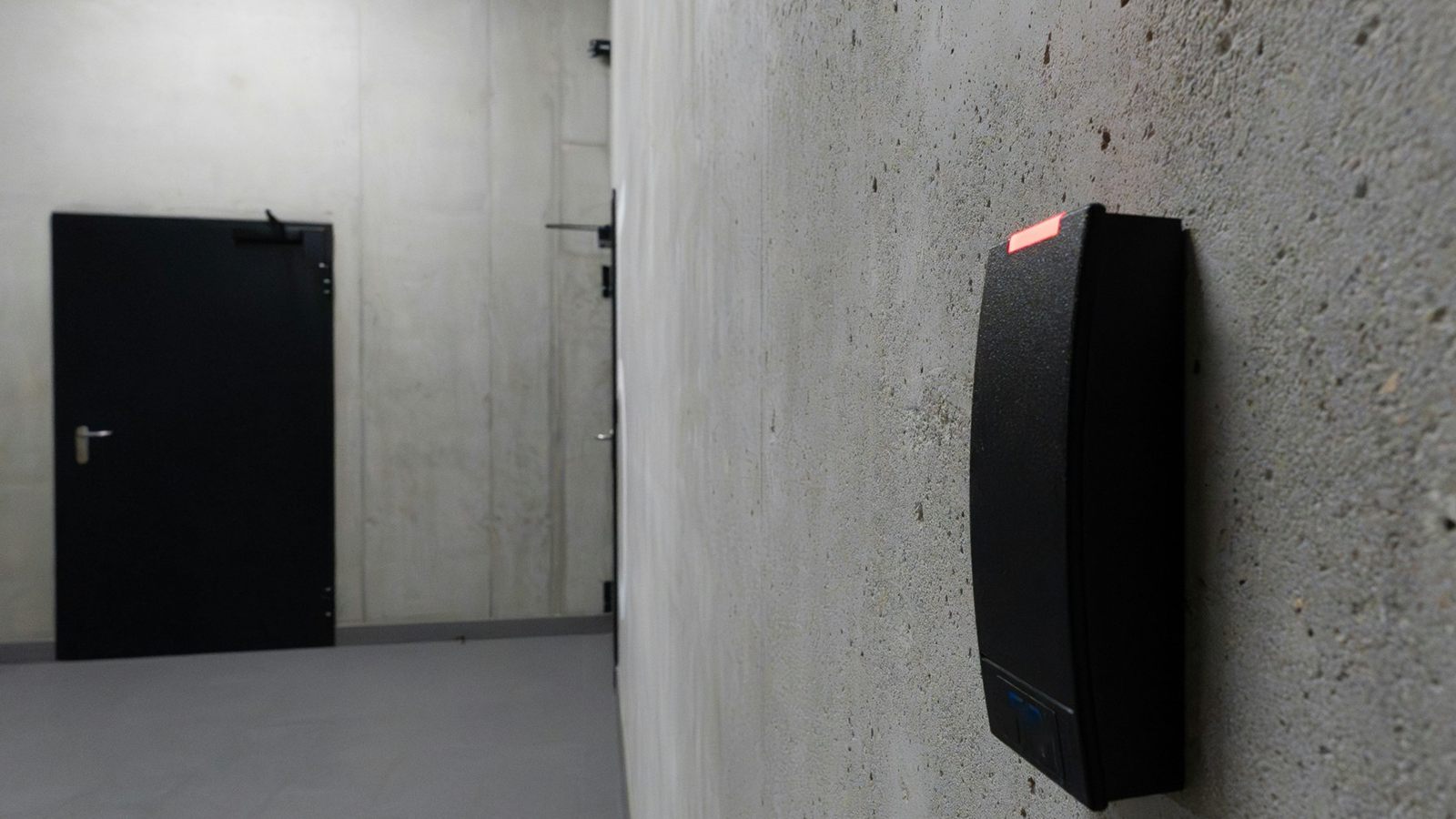 A wall-mounted access reader faces a closed black door across a concrete corridor.