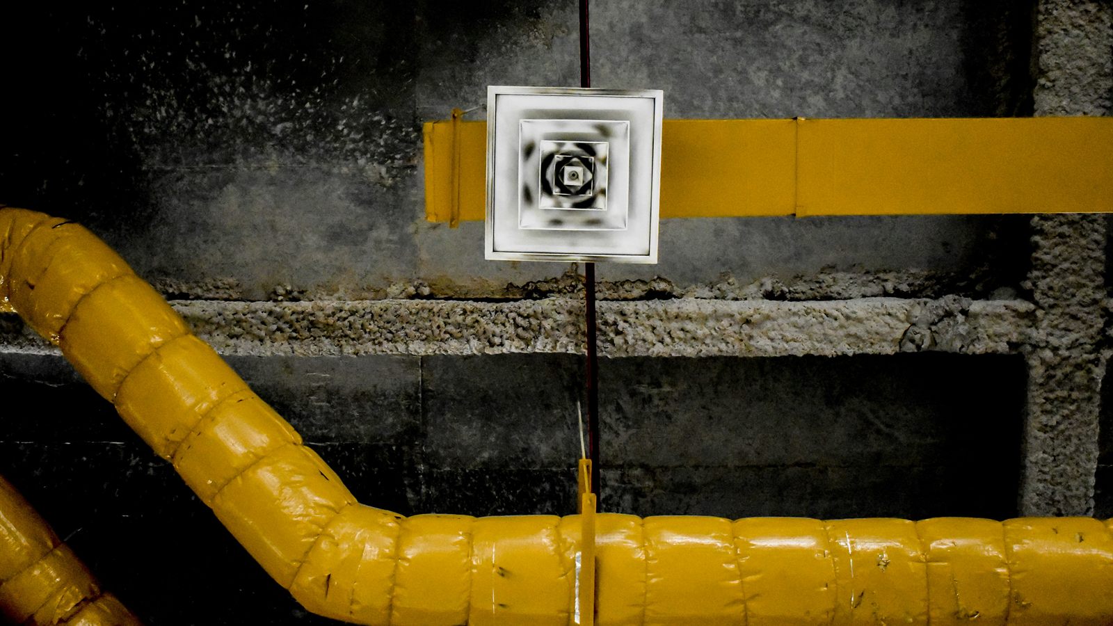 A square metal air vent sits between insulated yellow ducts in a mechanical room.