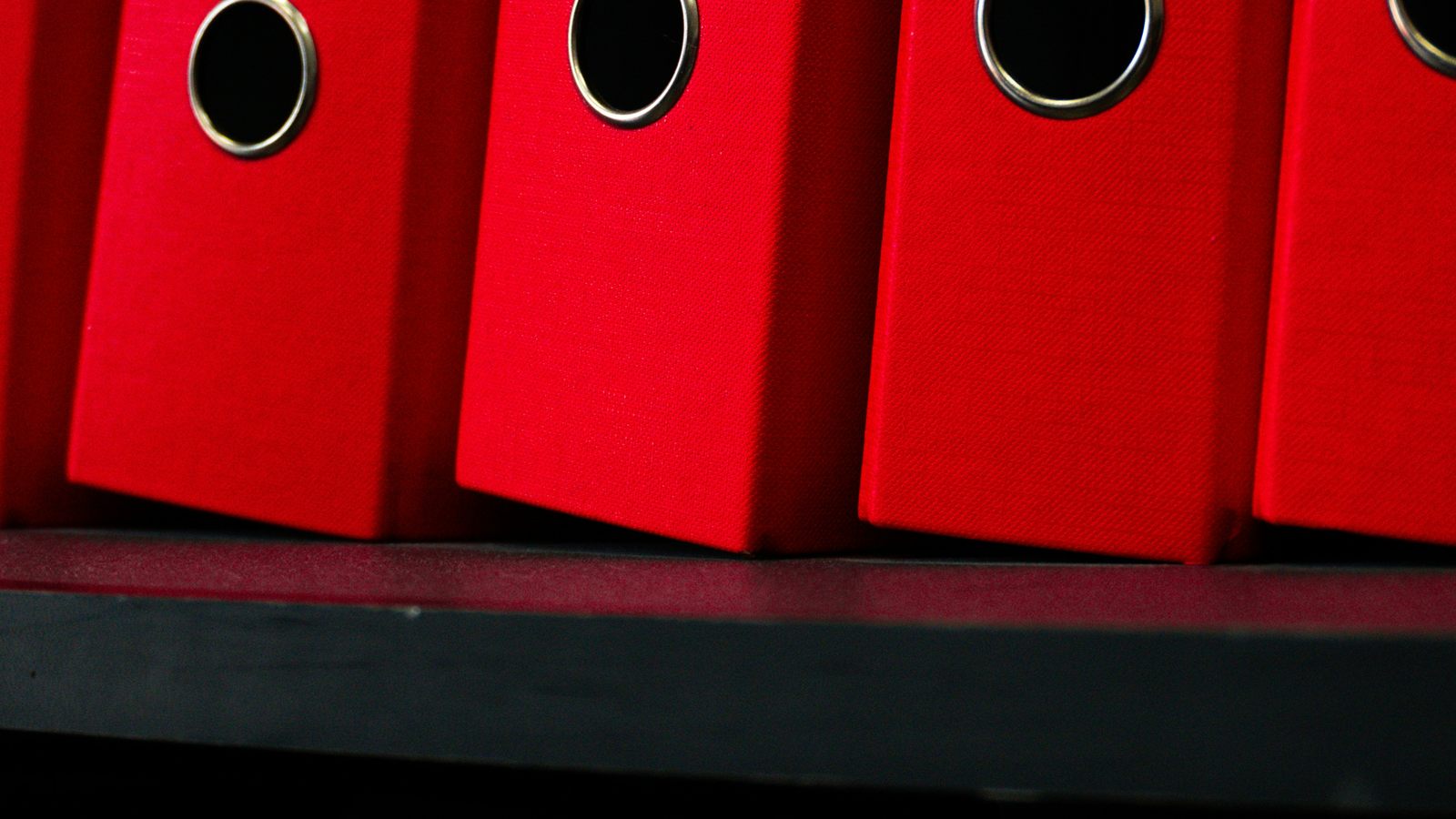 Red ring binders with black finger holes on a shelf.